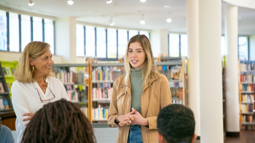 A woman listening to another woman while standing in a library