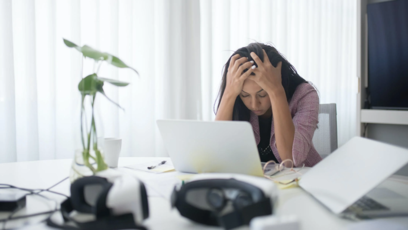 A tired woman is holding her head with both hands in front of a laptop