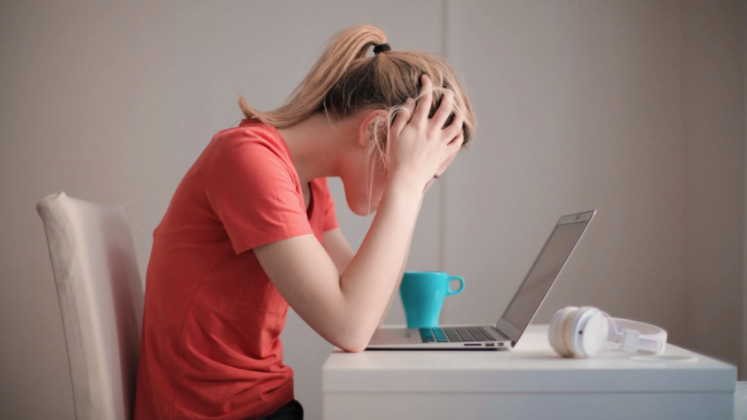 A stressed woman is holding her head in front of a laptop.